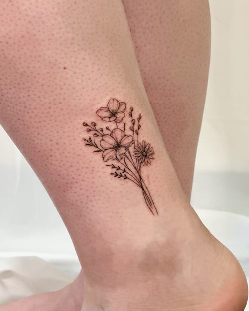 A delicate fine line flower bouquet tattoo on the ankle, featuring a minimalist black and grey design — by Melanie Benoit, los angeles