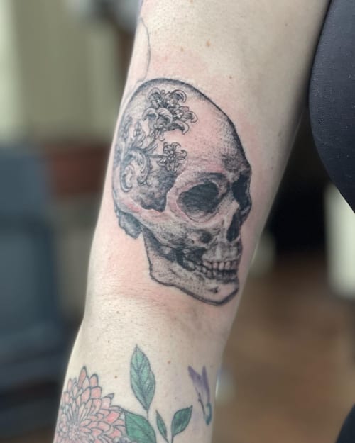 A black and grey realistic skull with floral design on the upper arm, featuring detailed shading and ornate elements — by Sandra Price, los angeles
