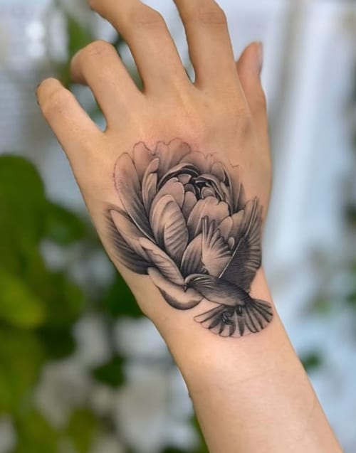 A black and grey realism tattoo on the hand featuring a detailed flower and a bird in flight — by otaybeatdown, los angeles