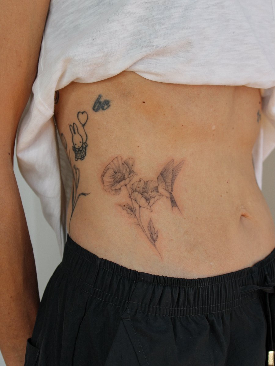 A delicate black and grey fine line tattoo of flowers and a hummingbird on the rib — by Melanie Benoit, los angeles