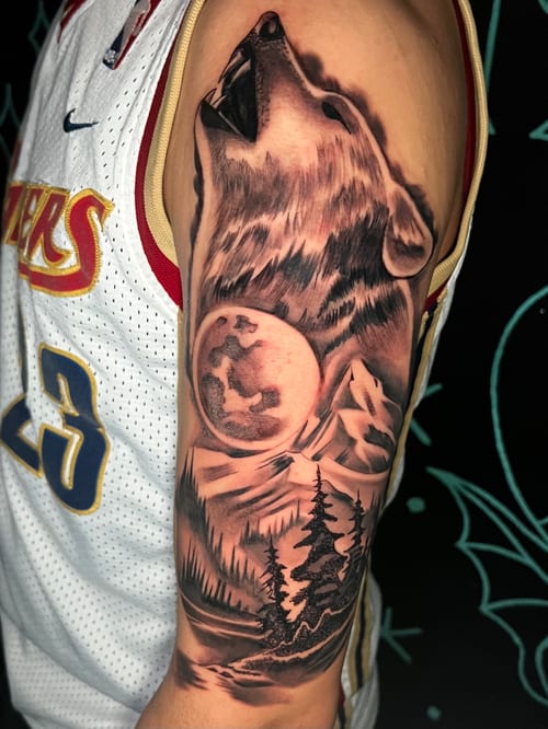 A realistic black and grey tattoo on the upper arm depicting a wolf howling at the moon with a forest landscape below — by Josue Bravo, los angeles