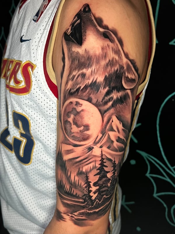 A realistic black and grey tattoo on the upper arm depicting a wolf howling at the moon with a forest landscape below — by Josue Bravo, los angeles