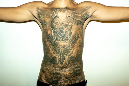 A detailed black and grey tattoo covering the full back, depicting an angel with outstretched wings above an ocean scene with ships, rendered in a realistic style — by Humilde tattoos, los angeles