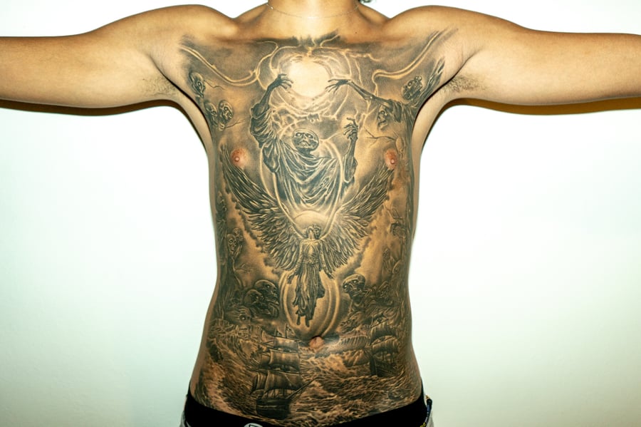 A detailed black and grey tattoo covering the full back, depicting an angel with outstretched wings above an ocean scene with ships, rendered in a realistic style — by Humilde tattoos, los angeles