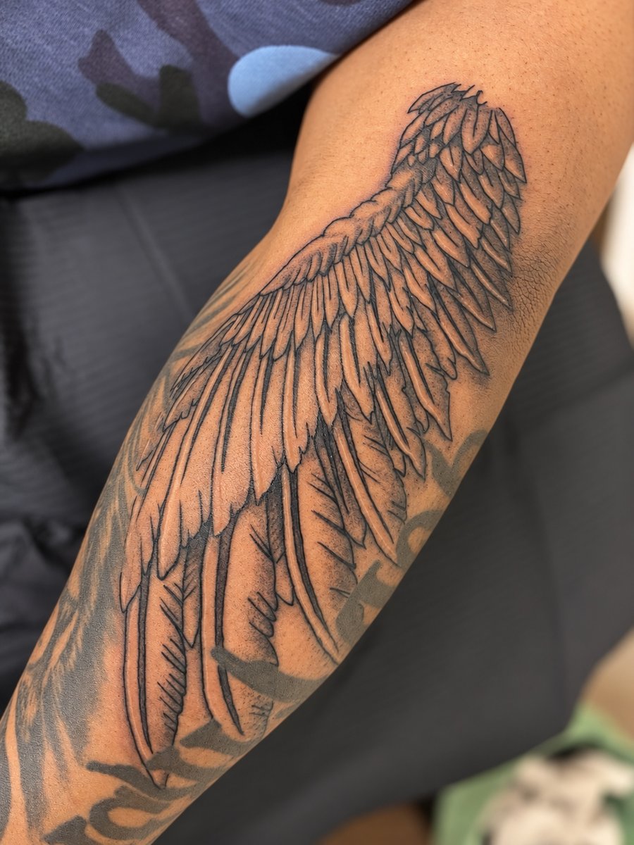 A detailed black and grey angel wing tattoo on the forearm with fine linework and delicate shading — by HEAVY INK, los angeles