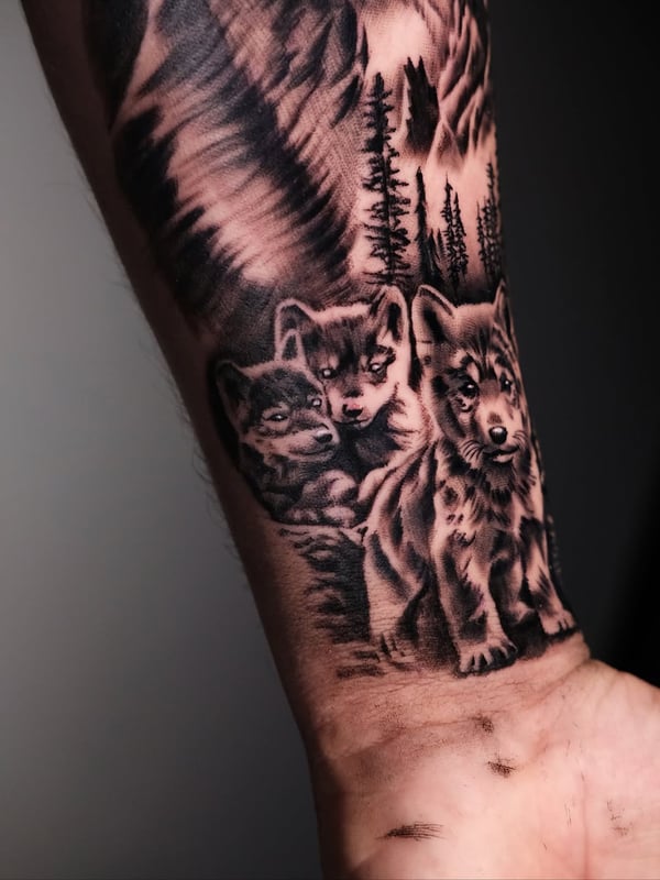 A detailed black and grey tattoo of wolf cubs in a forest setting on the forearm, showcasing realistic fur texture and shading — by Johnny Ayala, fontana, ca