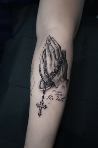 A realistic black and grey tattoo of praying hands holding a rosary with a cross, accompanied by a religious quote on the forearm — by Jian Liang, los angeles