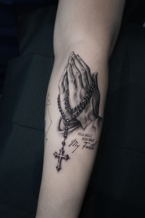 A realistic black and grey tattoo of praying hands holding a rosary with a cross, accompanied by a religious quote on the forearm — by Jian Liang, los angeles