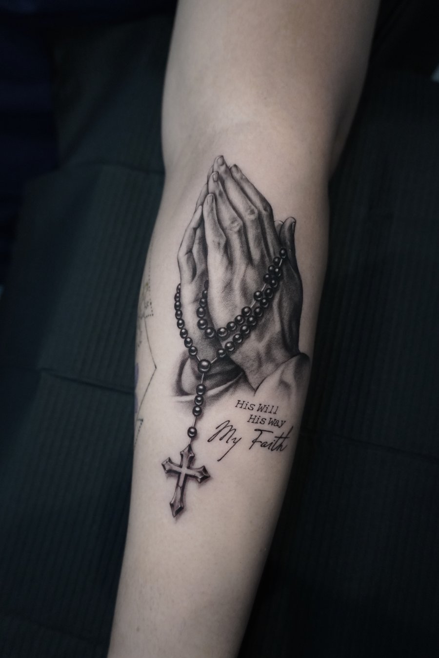 A realistic black and grey tattoo of praying hands holding a rosary with a cross, accompanied by a religious quote on the forearm — by Jian Liang, los angeles