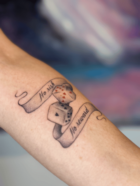 black and grey dice with card suit pips and a banner reading no risk no reward, forearm — by Samantha Wilks, los angeles