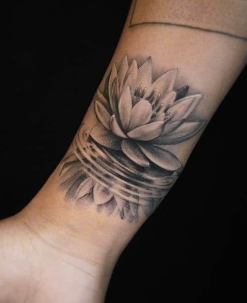 A black and grey realism tattoo of a lotus flower floating on water, located on the forearm with detailed shading — by Jian Liang, los angeles