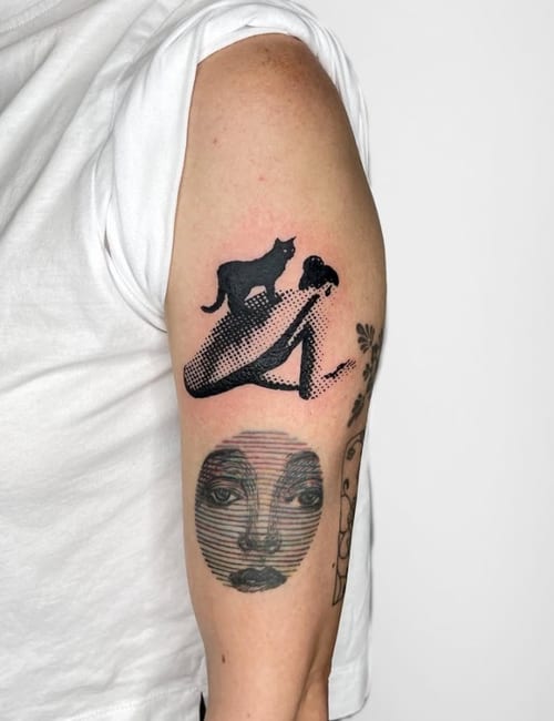 A black and grey illustrative tattoo on the upper arm featuring a stylized face and a cat with bold blackwork elements — by Level tattoo, los angeles