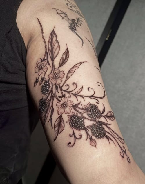 A delicate black and grey fine line floral tattoo on the upper arm, featuring intricate flowers and leaves with detailed linework — by Shelby Bianco, los angeles