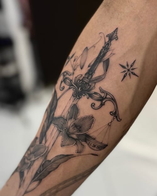 A black and grey fine line tattoo on the forearm featuring an ornate sword intertwined with flowers and a small date inscription — by otaybeatdown, los angeles