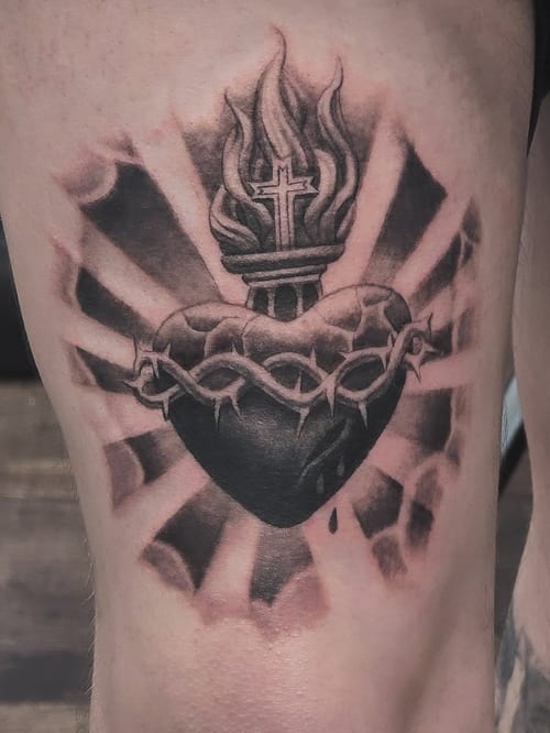 A bold black and grey sacred heart tattoo on the thigh, featuring flames and a cross, symbolizing religious devotion — by Miguel ANGELO, los angeles