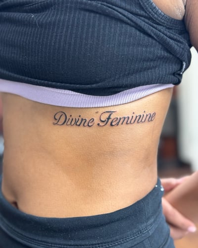 A minimalist black and grey script lettering tattoo on the rib reading 'Divine Feminine' — by Sandra Price, los angeles