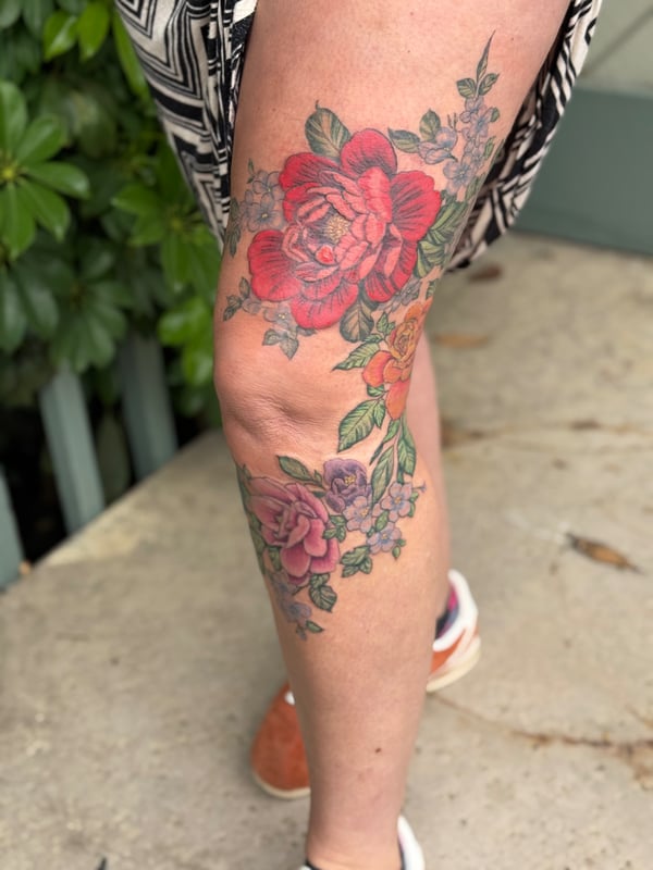 A colorful floral bouquet tattoo on the leg featuring roses and other flowers, with a delicate and romantic style — by Sandra Price, ventura, ca
