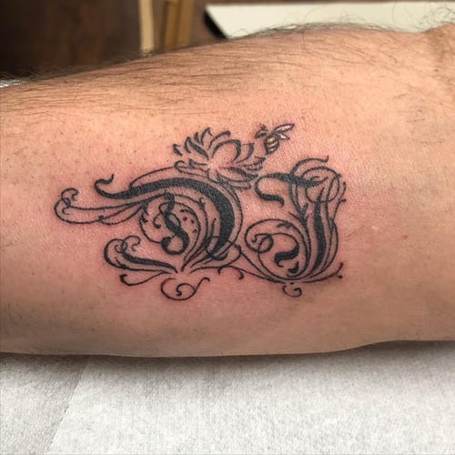 An ornate black and grey tattoo of initials with decorative elements on the forearm — by Sandra Price, los angeles