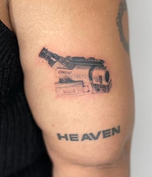 A black and grey minimalist tattoo of a camera on the upper arm with the word 'HEAVEN' in bold script below it — by Level tattoo, los angeles