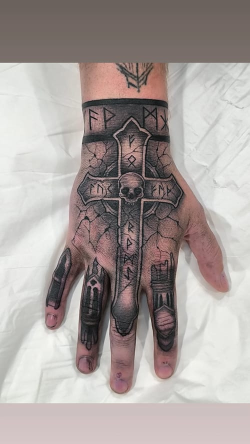 A black and grey cross tattoo on the hand featuring a skull and runes — by Samuel Connelly, los angeles