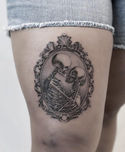 A black and grey tattoo of two skeletons embracing, framed in an ornate oval design on the thigh, with realistic shading and detail — by Jian Liang, los angeles