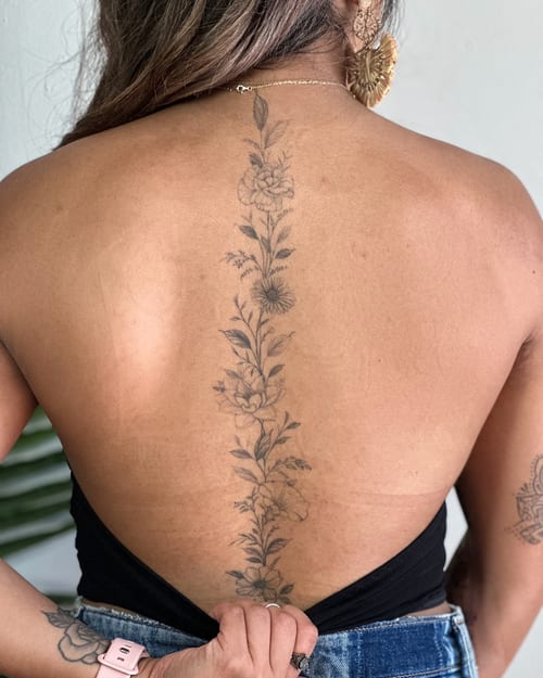 A delicate black and grey floral tattoo running vertically along the spine, featuring fine line work and intricate flower details — by Melanie Benoit, los angeles