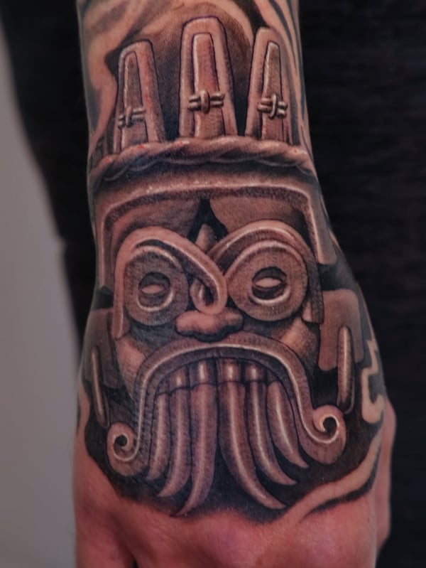 A bold black and grey Aztec mask tattoo on the hand with detailed shading and graphic elements — by Johnny Ayala, fontana, ca