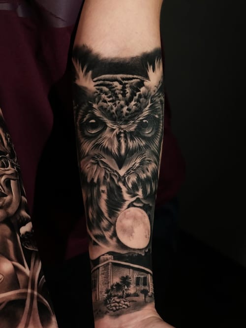 A realistic black and grey owl with a moon on the forearm, featuring detailed shading and texture — by Johnny Ayala, los angeles