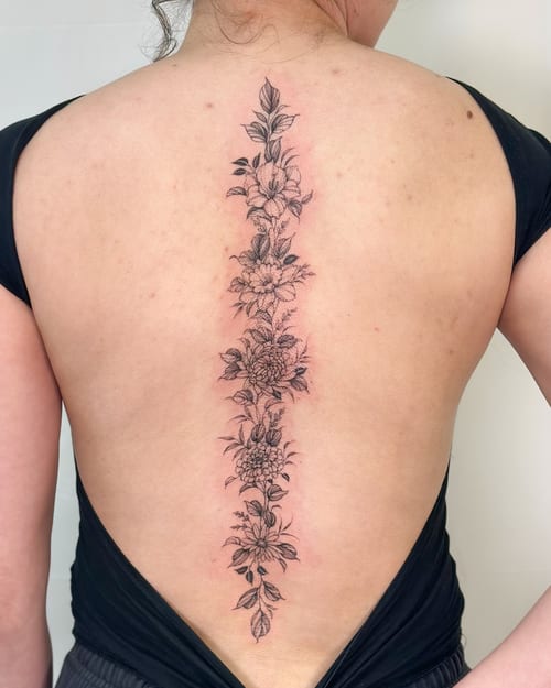 A delicate black and grey floral arrangement tattoo running vertically along the spine, featuring fine line work and intricate details — by Melanie Benoit, los angeles