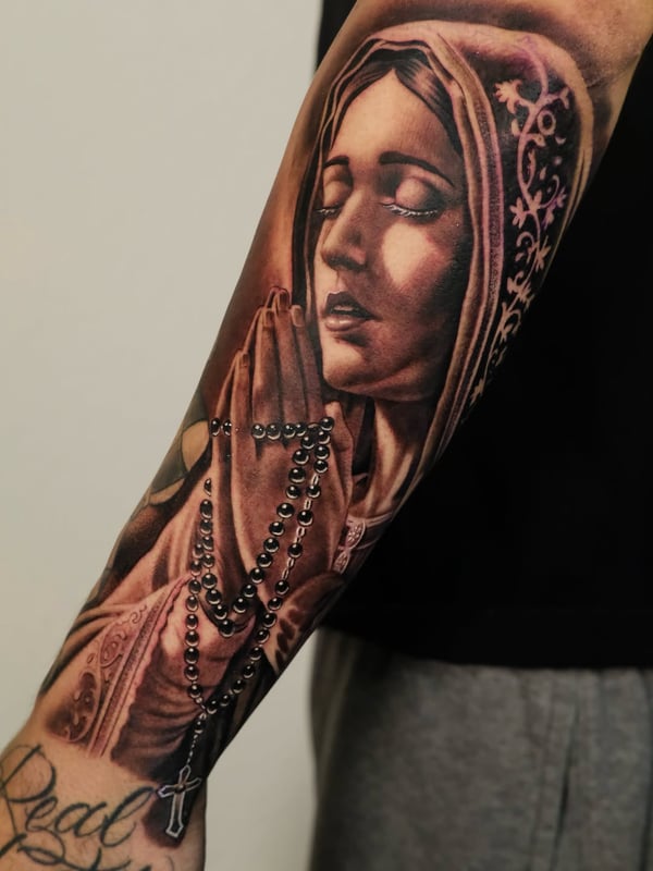 A realistic black and grey tattoo of a praying woman with a rosary on the forearm, featuring detailed shading and spiritual elements — by Johnny Ayala, fontana, ca