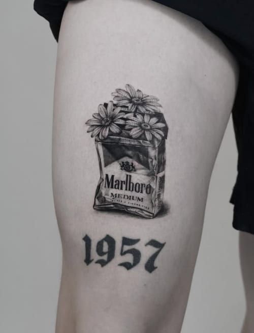 A black and grey tattoo of a Marlboro cigarette pack with flowers emerging from it, accompanied by the year '1957' in bold script on the thigh — by Jian Liang, los angeles