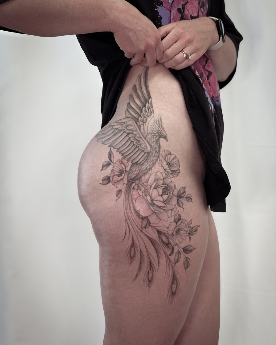 A detailed black and grey phoenix with floral elements on the thigh, featuring delicate and ornate linework — by Melanie Benoit, los angeles