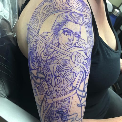 A bold linework tattoo of a warrior woman with a sword on the upper arm, featuring intricate Celtic patterns — by Sandra Price, los angeles