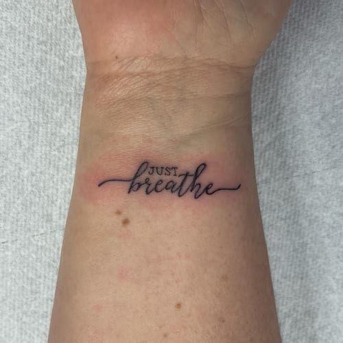 A minimalist black script lettering tattoo on the wrist with the phrase 'Just breathe' — by Sandra Price, los angeles