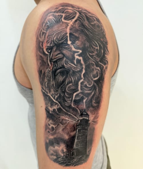 A black and grey realism tattoo on the upper arm depicting an intense portrait of a bearded man with lightning and a lighthouse in the background — by 钟逸政, los angeles