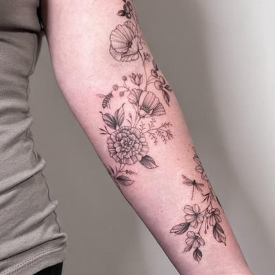A delicate black and grey floral arrangement with a bee and dragonfly on the forearm, featuring fine line work and botanical elements — by Melanie Benoit, los angeles