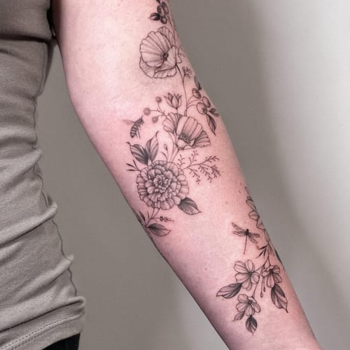 A delicate black and grey floral arrangement with a bee and dragonfly on the forearm, featuring fine line work and botanical elements — by Melanie Benoit, los angeles