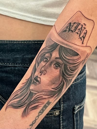 A black and grey portrait of a woman wearing a cowboy hat on the forearm, with detailed shading and script above the hat — by Josue Bravo, los angeles