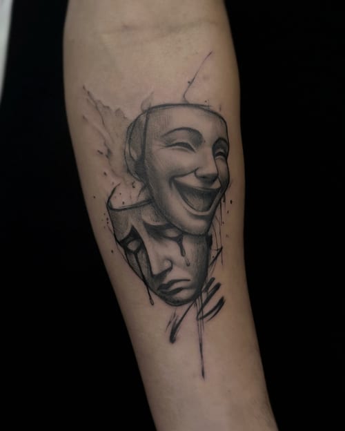 A black and grey tattoo of theatrical masks on the forearm, depicting one smiling and one sad face with realistic shading and detail — by Jian Liang, los angeles
