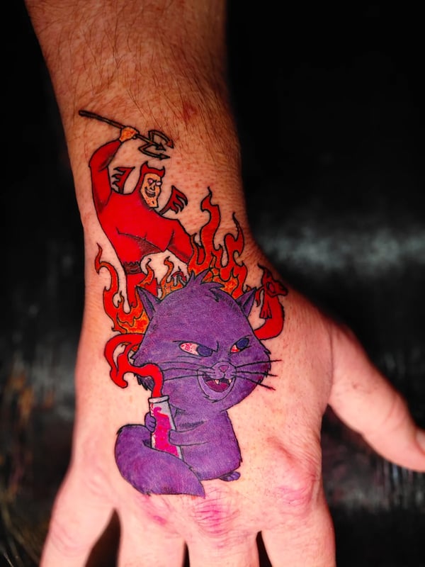 A colorful illustrative tattoo on the hand featuring a purple cat with a mischievous expression and a red demon-like figure in the background surrounded by flames — by anoetic.designs.ultd, redland…