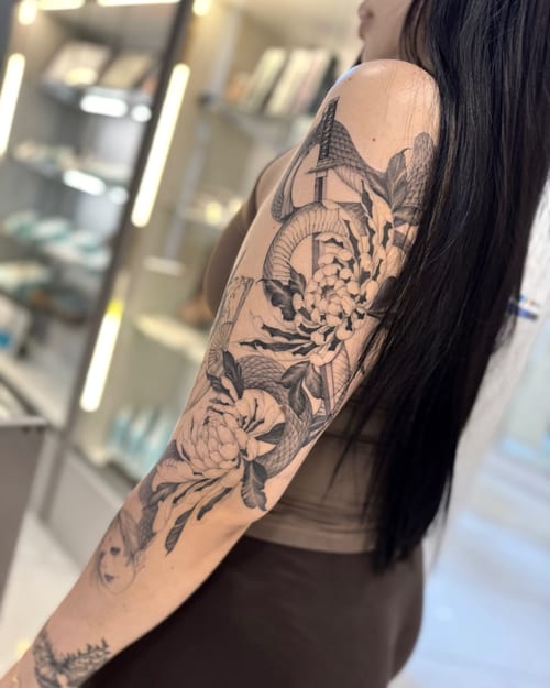 A black and grey floral tattoo on the arm featuring detailed flowers and leaves in a blackwork style — by otaybeatdown, los angeles