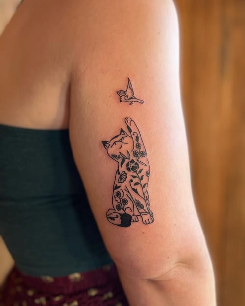 A whimsical black and grey tattoo of a floral-patterned cat reaching for a bird on the upper arm — by Dylan Aiello, los angeles