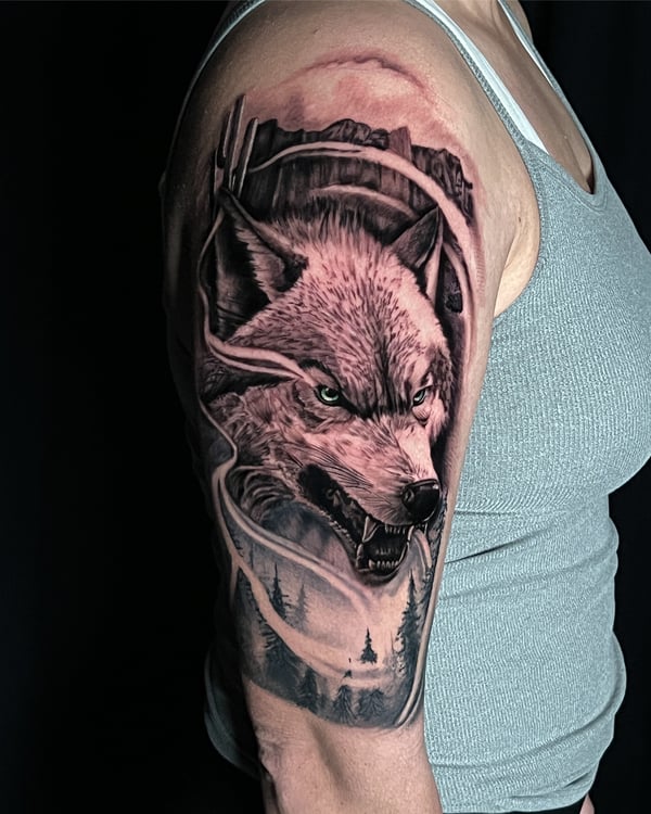 A detailed black and grey realism tattoo of a wolf with a forest and mountain background on the upper arm — by Travis Ross, inglewood, ca