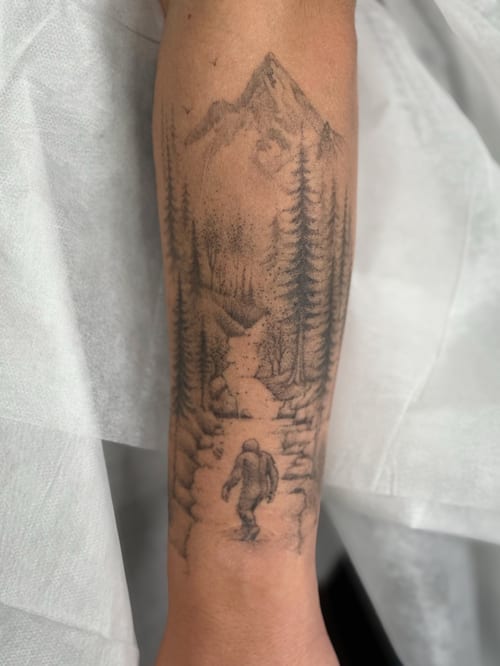 A black and grey tattoo of Bigfoot walking through a forest with mountains in the background on the forearm — by Melanie Benoit, los angeles