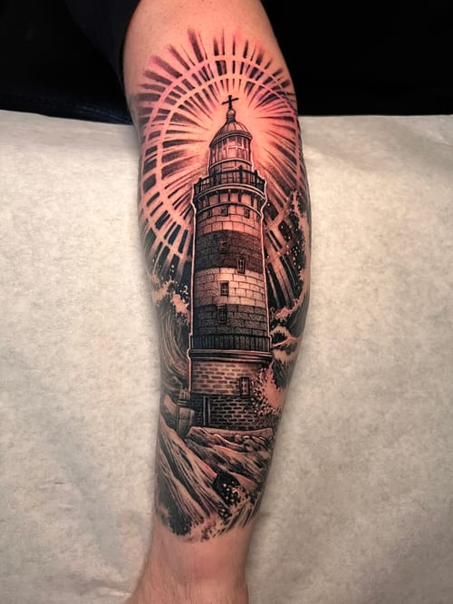 A detailed black and grey realism tattoo of a lighthouse on the forearm, with waves and a radiant light effect in the background, accented with subtle color highlights — by imindisoul, los angeles