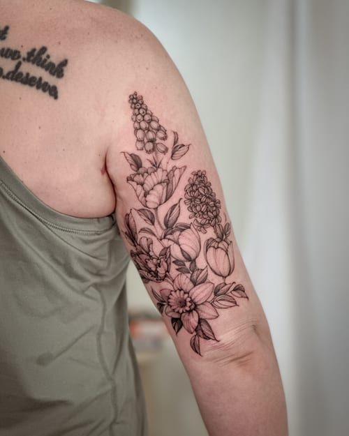 A delicate black and grey floral bouquet tattoo on the upper arm, featuring various flowers in a clean blackwork style — by Melanie Benoit, los angeles