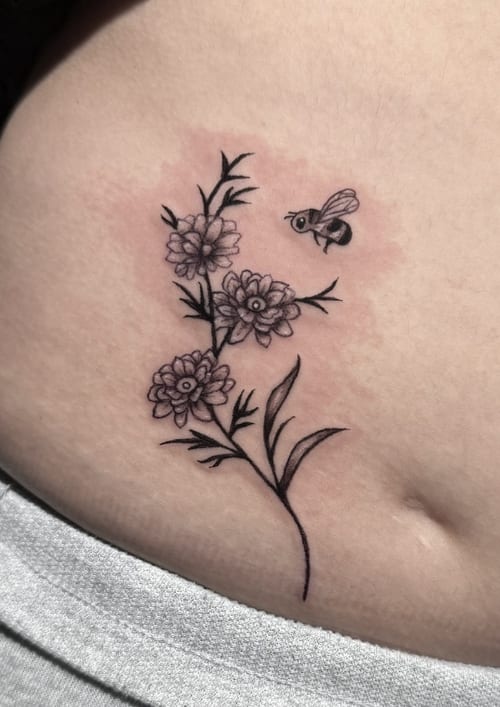 A fine line black and grey tattoo of flowers with a bee on the rib, featuring delicate linework and shading — by Shelby Bianco, los angeles