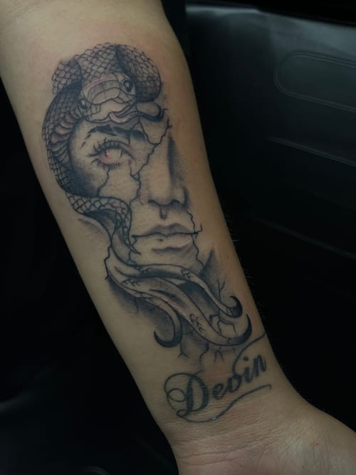 A black and grey surrealistic tattoo on the forearm featuring a snake intertwined with a fragmented face and script below — by Mr._Folks, los angeles