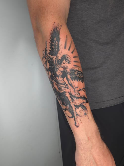 A detailed black and grey tattoo of an angel with wings on the forearm, featuring realistic shading and dynamic movement — by ashynucklestattoo, los angeles