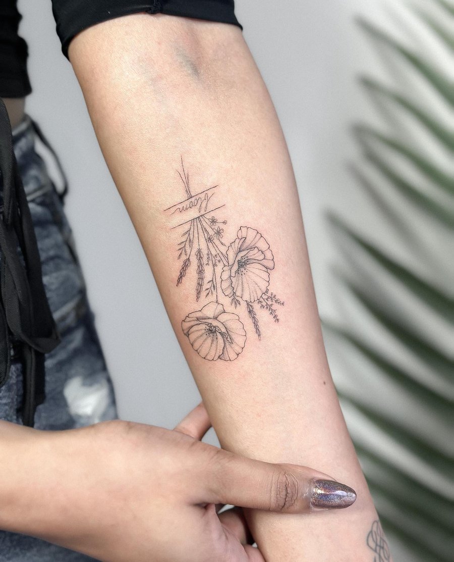 A delicate fine line tattoo of flowers with script on the forearm, done in black and grey — by Melanie Benoit, los angeles